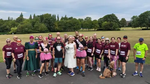 Christine Ball Sara Butler and Chris Hall standing in the middle of a group of about 27 people and a brown dog. Most are wearing maroon Low Fell Running Club t-shirts but a man and woman beside the couple are wearing black t-shirts with pink tutus and are holding pink bouquets of flowers. A woman on the left is wearing an 80s-style green bridesmaid dress with puffy sleeves and a gold band around the waist. To go with her wedding dress, Butler is wearing blue running trainers and Hall is wearing teal shorts and trainers along with his tuxedo t-shirt. They are standing in front of a large grassy field which has a row of trees at the back. Everyone is smiling.