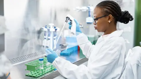 A scientist wears a white lab coat and works with test tubes in a laboratory. She is wearing glasses and has braided hair tied into a bun.