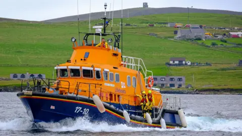 A bright orange lifeboat sailing through a narrow stretch of water. There are steep green hills in the background, sparsely populated by houses. Emergency workers are on the vessel wearing protective equipment.
