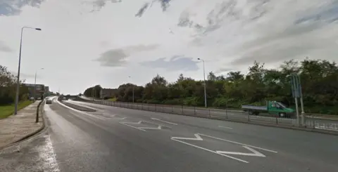 A street view image of the road, which is nearly empty. The image shows the road running into two directions, divided by a metal barrier in the middle.