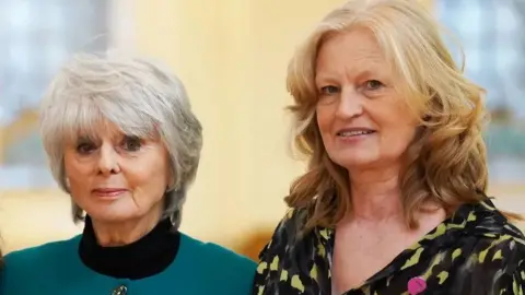 PA Media Diana Parkes stands to the left of the frame. She has short, grey hair and wears a black turtle neck jumper, with a teal blue collarless jacket with gold detailed buttons. She looks directly at the camera and is standing next to her friend, Hettie Barkworth-Nanton, who has mid-length, wavy, ginger hair and wears a V-neck yellow and black abstract printed blouse