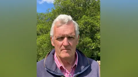 Peter Smith A head and shoulders image of Peter Smith. He has white hair and is wearing a red and white checked shirt with a navy fleece gilet over the top. He is frowning slightly. He is standing in front of a tree and blue sky can be seen beyond.