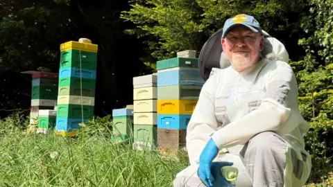 Beekeeper Ashley Galley wears a protective white suit next to series of coloured hives.