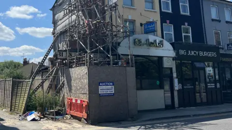 Scaffolding in Aylesbury that was put up in 2018. It is against the end building of a row of terraced businesses, which includes burger and Indian restaurants. The side of the building is being propped up with big metal joists that are angled into the ground, and there is tarpaulin across the brickwork. There is blue sky behind the building and white fluffy clouds, and there is a road in front of the scaffolding.