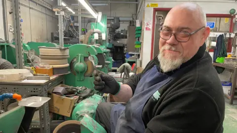 BBC / Jack Hadaway-Weller A smiling man wearing glasses sits at a machine in a workshop. He is holding medical tuning fork and is wearing black gloves.
