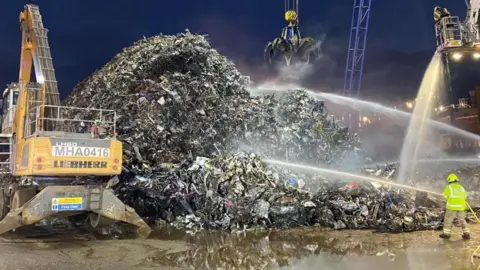 Firefighters attending an incident at at scrap metal yard at in the Port of Tilbury, it shows a pile of scrap metal, a fire fighter aiming a hose at the pile and heavy machinery