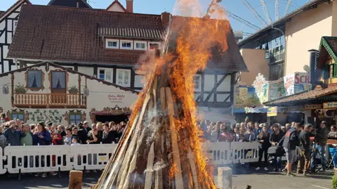 A town square with crowds behind a barrier with a large pile of wood on fire in the middle