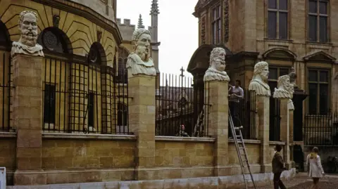 Oxfordshire County Council Michael Black is pictured sculpting the head figurines known as the Emperor's Heads mounted on pillars outside the Sheldonian Theatre in Oxford. A man can be seen at the foot of the ladder watching the sculptor at work.