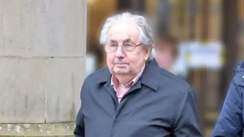 Spindrift A man with grey hair and glasses, wearing a blue overcoat and pink shirt leaves a court building