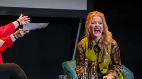 Bradford Literature Festival A woman on stage with a dark background behind her. She is mid-laugh. She wears a green waistcoat, chequered shirt and has long blonde hair.