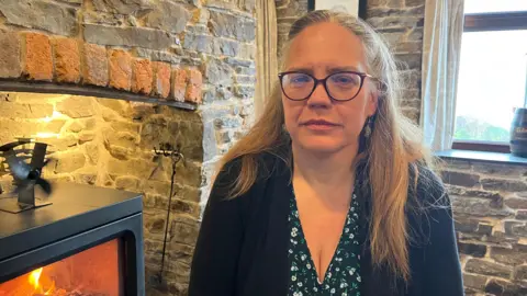 Sproson-Jones is photographed looking into the camera with a serious expression. She is wearing a black blouse with white and green flowers, and a black cardigan. Her hair is long and blonde, and is tied back in a half-up hairstyle. She's also wearing a pair of dark-framed glasses. She's sat next to a black log burner, which is lit and has flames inside. The walls of the pub and fireplace are exposed grey stone, with a window behind her. 
