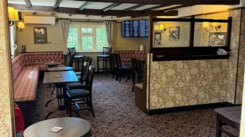 Neil Ainsworth The inside of a pub with dark and some partially red seats and tables on the left of the photo. There are lights on the walls, a window is around the centre of the photo at the back and a partition is on the right.