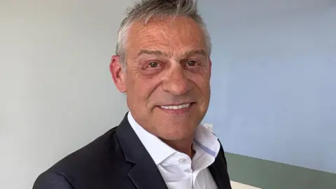 A man with grey hair is standing in front of a light coloured wall with a horizontal strip in a slightly darker shade. He is wearing a dark blazer over a white collared shirt.