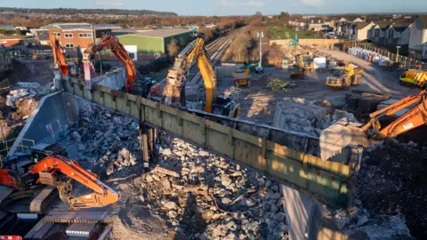 Diggers and other construction machinery are surrounded by rubble as a bridge is demolished over a railway track. It is a sunny winters day in a residential area.