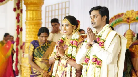 Janani Mohan and her husband in traditional Indian wedding clothing with garlands round their necks, stand with their hands in a prayer position in front of them, surrounded by guests celebrating their wedding