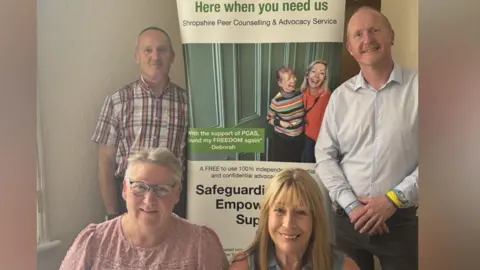 The picture show four people with a big promotional sign for PCAS. The man in the top left is wearing a red and white chequered shirt. The man in the top right is wearing a light blue shirt, with a yellow wristband and black trousers. The woman in the front right has blonde long hair and wears a blue vest. The woman in the front left has a pink t-shirt and is holding an appeal letter. She also has blue glasses and grey hair.