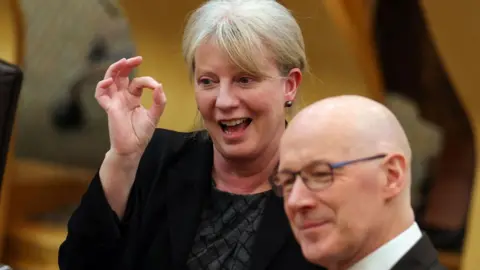 Getty Images Shona Robison, a woman with blonde hair tied back is holding, is holding her fore finger and thumb together in the OK sign. She is wearing a black jacket and black top. John Swinney, a bald man with glasses and wearing a black jacket, whit shirt and purple tie, is sitting behind her