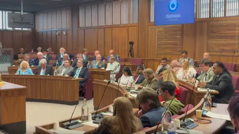 LDRS Oldham Council's chamber: a wood-panelled room with red seats and wooden desks in a u-shape around a central podium. There are people sitting in most of the seats. At the back is a screen with the Oldham Council logo.