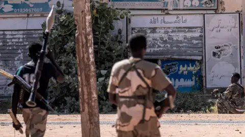 AFP Three Sudanese men in military uniform all pictured with their back to the camera and all with guns, the first on the left also holding an RPG pointed upwards. They all facing a building with Arabic inscriptions