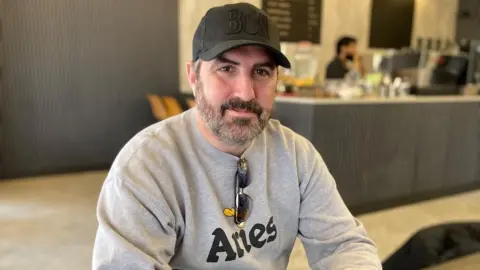 Alex Pope/BBC A man sitting in a coffee shop, with a cap on his head, wearing a grey jumper and glasses round his neck. The coffee bar is blurred behind him.