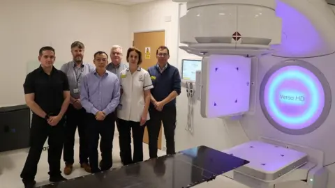 University Hospital Coventry and Warwickshire NHS Trust A group standing next to a radiotherapy machine in a hospital environment. The walls and machine are white but a circular section of the machine is glowing purple and blue and has the words Versa HD on a screen. The group of five men and one woman are posed facing the camera.