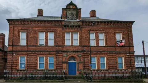 A two-storey red brick building with a central blue door and decorative stonework. The building has a “sold” sign displayed in an upper window.