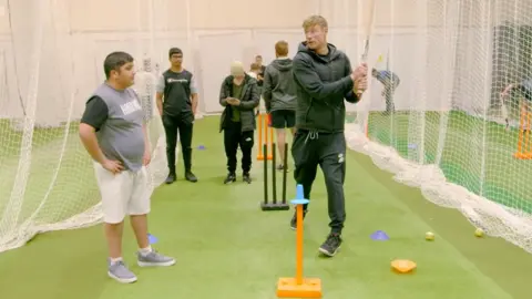 Freddie Flintoff is wearing a dark coloured tracksuit and is holding a cricket bat in cricket nets and is speaking to Umar Mahmood who is wearing white shorts and a grey top with grey pumps.