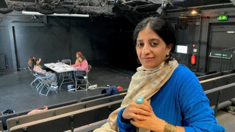 A woman wearing a blue jumper and yellow scarf with four people sitting around a table in the background