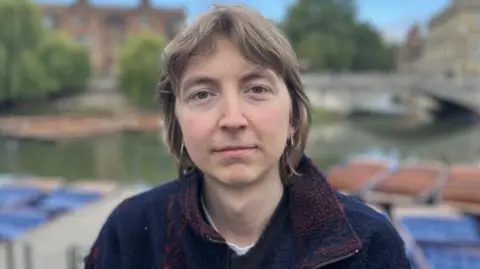Phil Shepka/BBC A head-and-shoulders picture of William Ridgeon. He is standing alone in Cambridge city centre, with the river behind him. He has shoulder-length brown hair and is wearing gold earrings. He his wearing a blue zip-up top.