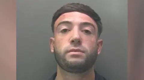 Custody image of Dominic Bevilacqua - he is looking into the camera with his chin raised. he has black hair and a short stubble beard