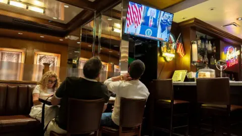 Customers at a bar in New York watch Trump's address on a screen on 1 April