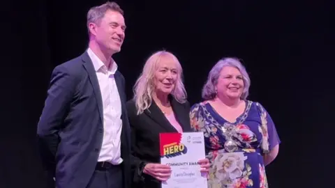 Three people stood together against a black background. One lady in the middle holds a "Community Award" piece of paper, and a man and woman stand on either side of her and smile.