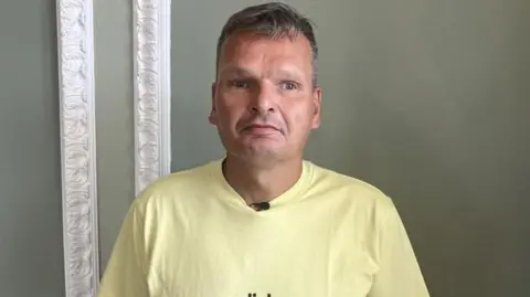 A man, William Fleming, in a yellow t-shirt stood in front of a green wall with white moulding. He has a serious expression.