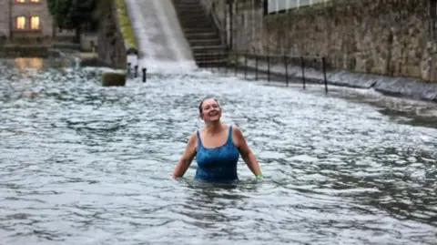 Louise Snelson Louise Snelson wearing a blue swimming costume stands in waist-deep water near a slipway. She is looking into the distance and has a big smile on her face. She has a green watch on her left arm.