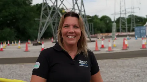 Hannah Karpel/BBC woman in the foreground wearing a black polo shirt with a “Morgan Sindall Infrastructure” logo, standing at an outdoor electrical infrastructure site and facing the camera. Behind the person are large steel electricity transmission structures, overhead power lines, and a gravel-covered work area. Numerous orange-and-white traffic cones mark pathways and restricted areas across the site. Trees line the background, and a blue site container is visible on the right. No bags or bins are visible.