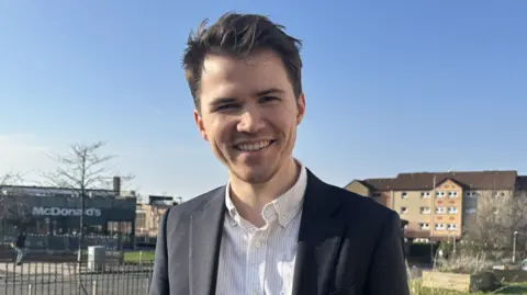 Gordon McKee is a young man, in a stripe shirt and blazer with short dark hair. He is smiling, standing outside the Braes, the McDonalds drive thru behind him.