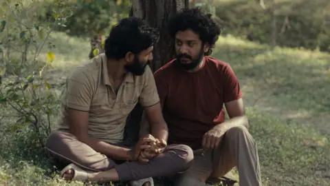 Lotus Visual Productions The image shows two men holding hands and talking to one another sitting by a tree in an open field.