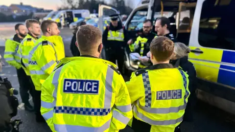 In the foreground a team of 10 police officers stand in a circle, wearing high visibility Jackets with POLICE written in white on the back. Their police van is behind them to the right. Blurred in the background you can see other teams getting into their vans. The lighting is subdued as it's taken at dawn.