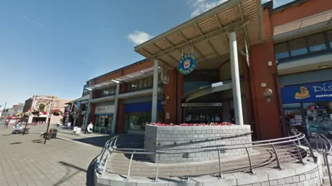 Barrow Market Hall pictured from the street outside. There is a grey stone planter containing red flowers in front of the hall, which is a red brick building with metal pillars holding up the roof. In the centre dangles a round blue sign with the words Barrow Market and a heraldic shield. Shop fronts span out from a central entrance.