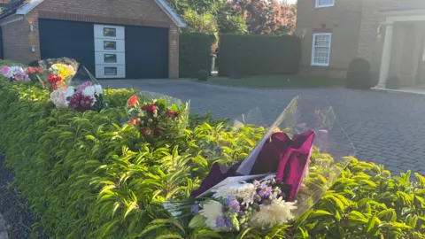 Holly Nichols/BBC Bunches of plastic-wrapped flowers pushed into a green hedge in front of a house in Carnoustie Drive in Great Denham. Behind the hedge is a garage and a corner of the detached brick-built house and its front entrance.