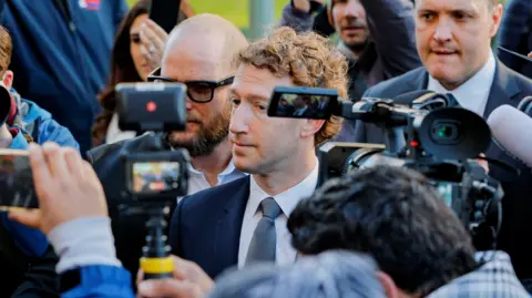 Reuters Meta owner and Facebook founder Mark Zuckerberg in a dark blue suit, white shirt and grey tie, stands surrounded by people and cameras, outside court during the case against Meta and Google.