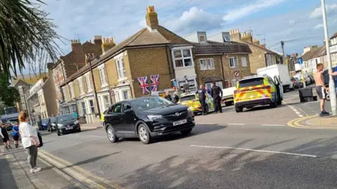 Danny Ashton-Keenan Road junction with police cars and uniformed officers. People are milling about. A number of civilian cars also visible. Union flags are seen hanging from a building.