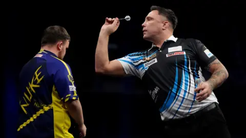 Gerwyn Price, pictured throwing a dart at a dartboard out of shot. He is wearing a black shirt with blue and white shards across it and a white collar. Luke Littler has his back to the camera, looking straight ahead. The background is black. 
