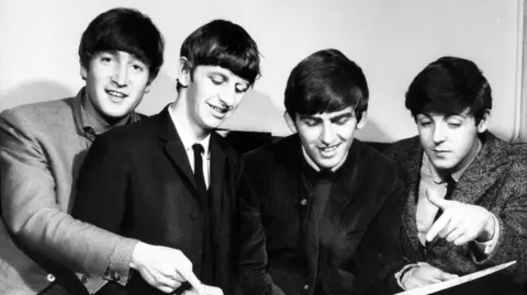 A black and white picture showing the Beatles, who smiling while looking at a large book. Sir Ringo, second from left, is holding a pen in his right hand.