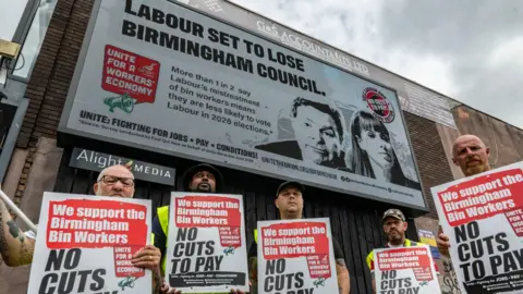 Getty Images Striking bin workers stand in front of billboards commissioned by the Unite trade union, holding placards in support of striking bin workers