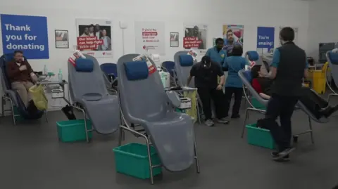 The blood-giving room in a hospital, with donors sitting in big chairs as they give blood. A number of the chairs are empty and the wall bears posters thanking people for donating.