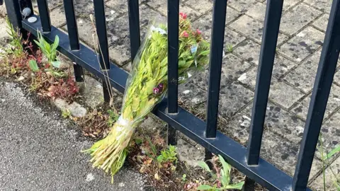A bouquet of flowers rests between metal fence posts outside a primary school. There are weeds growing beneath the fence, between the pavement and the block paving on the school grounds.