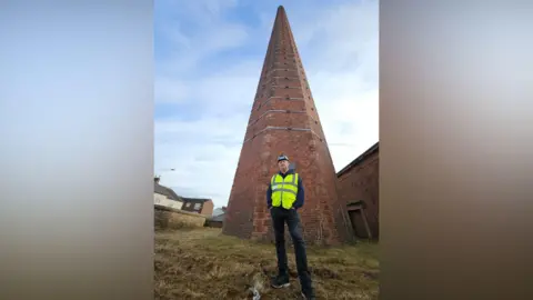 Rafferty Specialist Access Services' contracts director Carl Woodroffe standing at the bottom of Dixons Chimney in Carlisle. He is wearing a hard hat and yellow hi-vis vest, over a blue jacket and black jeans. He is looking down at the camera. Dixons Chimney is a red-brick structure with a hexagon shape. There are metal belts around the structure.