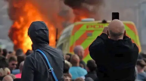 BBC Scene from the riots in Southport in July 2024. Two men are pictured from behind looking towards a police van and flames. One has the hood of his coat up, the other has a bald head and is pointing his mobile phone towards the scene. Between the two men and the fire are a crowd of people. 