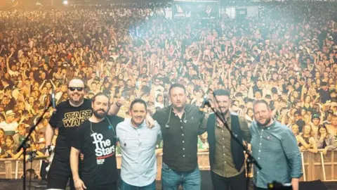 Oscar Bradley The band standing next to each other on stage with the crowd behind him.